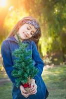 Linda chica joven de raza mixta sosteniendo un pequeño árbol de navidad al aire libre foto