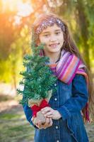 Linda chica joven de raza mixta sosteniendo un pequeño árbol de navidad al aire libre foto