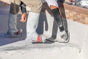 trabajador que usa zapatos con púas alisando el yeso húmedo de la piscina con una paleta foto