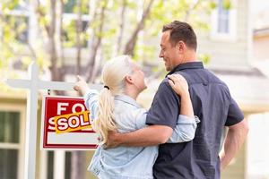 Caucasian Couple Facing and Pointing to Front of Sold Real Estate Sign and House. photo