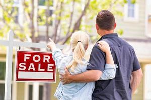 Pareja caucásica mirando y apuntando al frente de la casa y el cartel de venta de bienes raíces foto