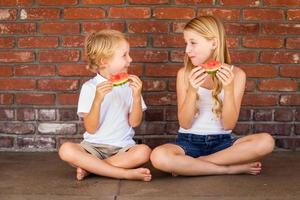 Cute Young Cuacasian Boy and Girl Eating Watermelon Against Brick Wall photo