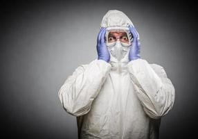 Man Holding Head With Hands Wearing HAZMAT Protective Clothing Against A Gray Background. photo