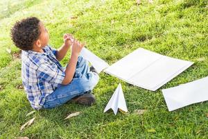 niño afroamericano y mexicano aprendiendo a doblar aviones de papel al aire libre en la hierba foto