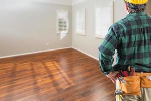 Contractor Wearing Toolbelt and Hard Hat Facing Empty Room with Hard Wood Floors photo