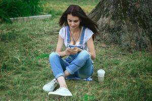 young charming brunette sits in the grass listening to music and looking at phone photo