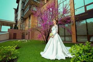 blonde bride in wedding dress and roses in her hands posing with sakura tree on background photo