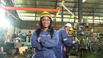 ritratto di professionale nero femmina lavoratore nel protettivo sicurezza uniforme e casco guardare a telecamera, braccia attraversato e sorridente, con ingegneri squadra dietro a sua nel un' metallo produzione fabbrica. video