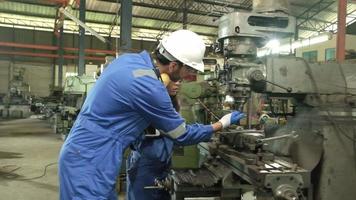 twee industrieel arbeiders in beschermend en veiligheid uniformen en harde hoeden, mannetje manager, en zwart collega werk met metaalwerk machines in fabricage fabriek. professioneel productie ingenieur team. video