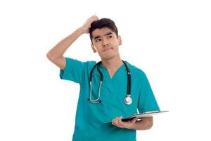 young funny brunette man doctor in blue uniform with stethoscope on his shoulders make notes and thinking isolated on white background photo