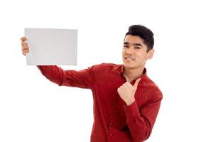 Un joven elegante con camiseta roja con una pancarta vacía mirando a la cámara y mostrando los pulgares hacia arriba aislado de fondo blanco foto