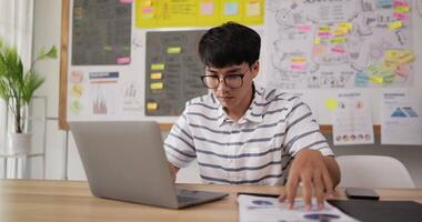 retrato de óculos de homem asiático digitando laptop e verificando documento enquanto está sentado na mesa do local de trabalho no escritório. cara jovem focado trabalhando no documento do projeto no escritório moderno. video