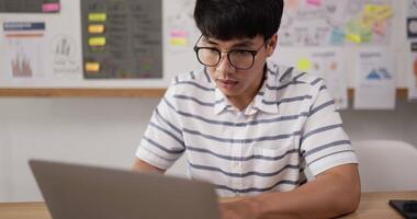 Closeup of Asian man glasses typing laptop and checking document while sitting at workplace desk in office. Focused young guy working on project document at modern office. video