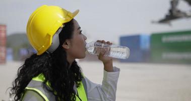 Pretty young Engineer woman wearing helmet and safety vest sitting and drinking water, She wipe sweat on face while working in the port, shipping container Logistic concept video