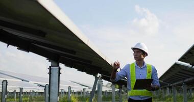 Handheld medium shot, front view of Asian young inspector engineer man wearing white helmet and hold report paper in hand walk to checking solar panel in solar farm video