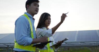 handhållen spårning skott, bakgrundsbelyst asiatisk ung inspektör ingenjör man och kvinna kollega gående mellan rad av sol- panel medan kontroll drift i sol- bruka video
