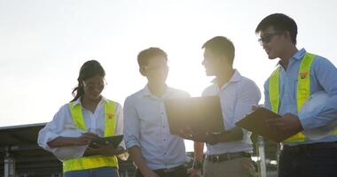 toma retroiluminada, un grupo de ingenieros asiáticos usa una computadora portátil y una tableta durante la operación de planificación y el panel solar fotovoltaico en la estación, los jóvenes ingenieros inspectores y las operaciones de planificación femenina video