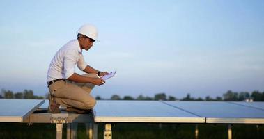 Seitenansicht des asiatischen jungen Inspektor-Ingenieurmannes mit weißem Schutzhelm, der sitzt, um den Betrieb des Solarmoduls in der Solarstation zu überprüfen video