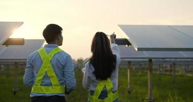 prise de vue à main levée, rétro-éclairé, vue arrière d'un jeune ingénieur inspecteur asiatique et d'une collègue marchant entre une rangée de panneaux solaires tout en vérifiant le fonctionnement de la station solaire