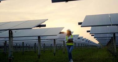 escena retroiluminada, joven inspectora asiática ingeniera con casco de protección sosteniendo papeleo en las manos caminando entre filas de paneles solares mientras revisa el funcionamiento en la estación solar por la noche video