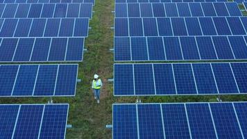 Aerial view from drone over Asian Young Inspector Engineer female wearing protection helmet hold paperwork walking between row of solar panel while checking operation in solar station in evening video