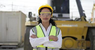portrait d'une belle femme ingénieur cheveux bouclés portant des lunettes, un casque et un gilet avec les bras croisés, sourire et regarder la caméra tout en travaillant dans le port du terminal à conteneurs video