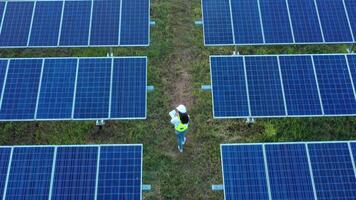 Aerial view from drone over Asian Young Inspector Engineer female wearing protection helmet hold paperwork walking between row of solar panel while checking operation in solar station in evening video