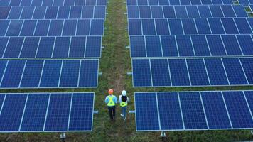 Aerial view from drone flying over Asian young inspector engineer man and woman wearing helmet and hold report paper in hand walk to checking solar panel in solar station video