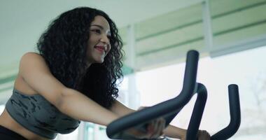 prise de vue au ralenti à la main, heureuse jeune femme asiatique machine à vélo spinning pour l'entraînement, belle femme aux cheveux bouclés exercice avec spin sur simulateur de vélo à air pour la santé cardiologique dans la salle de gym. video