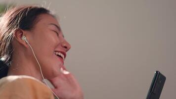 bella giovane asiatico donna seduta e godere per ascoltando musica e cantando a partire dal senza fili cuffie con smartphone nel vivente camera a casa, rilassante e stile di vita concetto. video