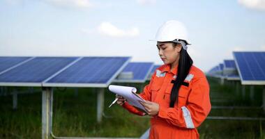 Handheld medium shot, Asian Young engineer woman wearing protection uniform and white helmet standing at front of solar panel to checking operation in paperwork while working in solar farm video