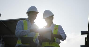 Slow motion shot, Backlit view Asian Young Inspector Engineer man and female colleague wearing white helmet checking operation in solar farm, They talking and smile, woman checking in paperwork video