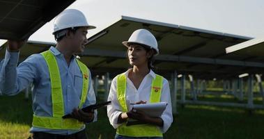 Slow motion shot, Asian Young Inspector Engineer man and female colleague wearing helmet standing and checking operation in solar farm, They are talking together and woman record to paperwork video