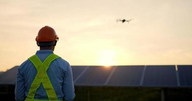 cena retroiluminada, homem jovem engenheiro inspetor asiático usando drone de controle de capacete de proteção voando sobre a linha do painel solar enquanto verifica a operação na estação solar à noite video