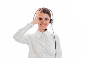 joven con camisa blanca y auriculares con micrófono sonriendo mantiene la mano cerca de los ojos y mirando a la cámara foto