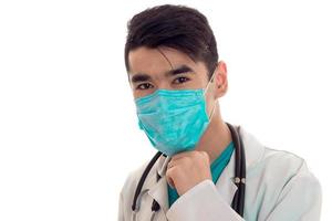 young brunette man doctor in white uniform and mask with stethoscope looking at the camera isolated in studio photo