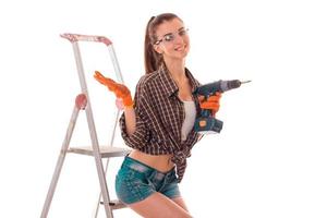 hermosa joven con gafas, pantalones cortos y camisa se para cerca de las escaleras y mantiene el taladro foto