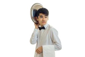 un camarero apuesto y divertido mira hacia otro lado y mantiene la bandeja del plato cerca de su cabeza aislada en el fondo blanco foto