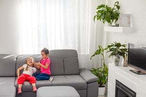 quédate en casa mantente a salvo. dos niños felices sentados en una silla acogedora relajándose jugando en una sala de estar blanca en el interior. hermanas divirtiéndose en casa foto