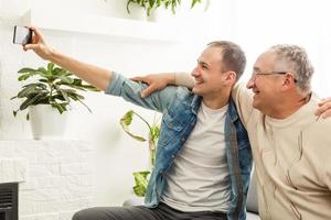 Happy moment. Cheerful young man taking a selfie with his upbeat elderly father waving at the camera and smiling pleasantly photo