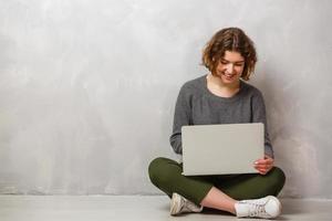 Portrait of satisfied female with beautiful smile enjoying watching movie in silver computer and sitting in lotus pose on the floor over grey wall photo