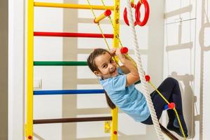 retrato de un niño feliz en un centro de entretenimiento de juegos. la niña sudorosa colgada de una suave barra horizontal. un hermoso bebé activo practica deportes. foto