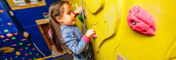 Little baby girl with funny hear style climbing vertical wall and man belaying her from below photo