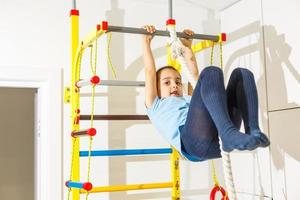Portrait of a happy child in a gaming entertainment center. The sweating little girl hanging on a soft horizontal bar. A beautiful active baby goes in for sports. photo