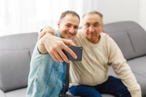 Happy moment. Cheerful young man taking a selfie with his upbeat elderly father waving at the camera and smiling pleasantly photo