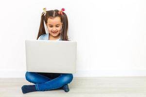 una niñita linda está sentada en el suelo con su laptop, usando anteojos, aislada en blanco foto