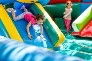 Child jumping on colorful playground trampoline. Kids jump in inflatable bounce castle on kindergarten birthday party Activity and play center for young child. Little girl playing outdoors in summer photo