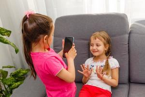 people, children, technology, friends and friendship concept - happy little girls sitting on sofa and taking selfie with smartphone at home. photo