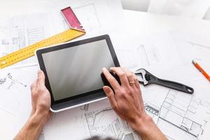 Man hand working with tablet and blueprint on architectural project at home while relaxing on couch. Back view of latin architect reading plan layout on digital tablet. photo