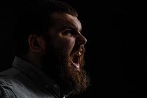 Naked bearded man angrily screams into a spray of water against a black background with copy space. Emotional portrait of a man like a barbarian. Toned image. side view close up view photo